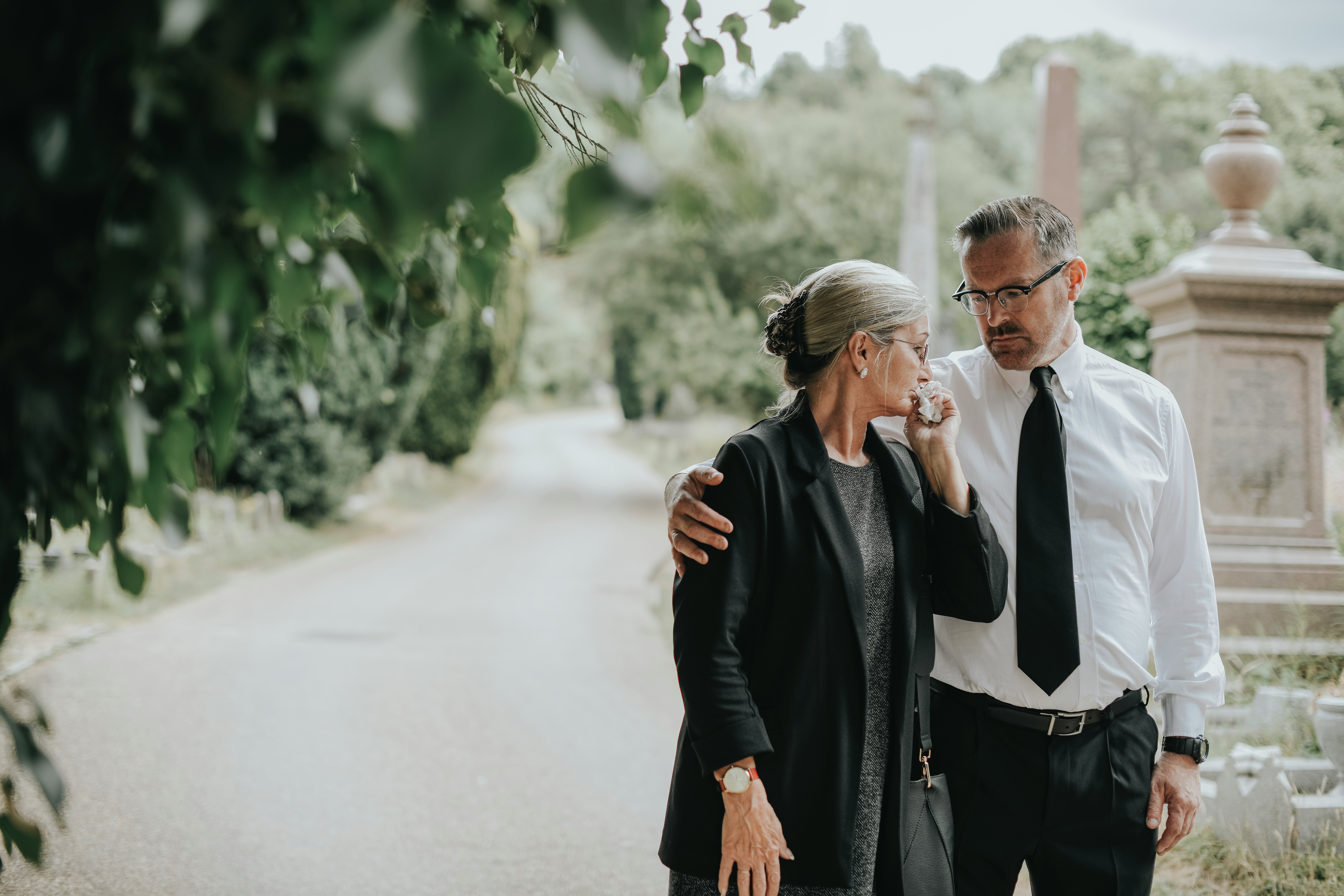 What To Do When Someone Dies Checklist | A Guide for California Families An older couple dressed in formal black clothing walk slowly along a quiet cemetery path, with the man gently holding the woman close as she wipes tears from her face, surrounded by greenery, stone monuments, and a soft, muted outdoor setting that conveys grief and comfort after a loss.