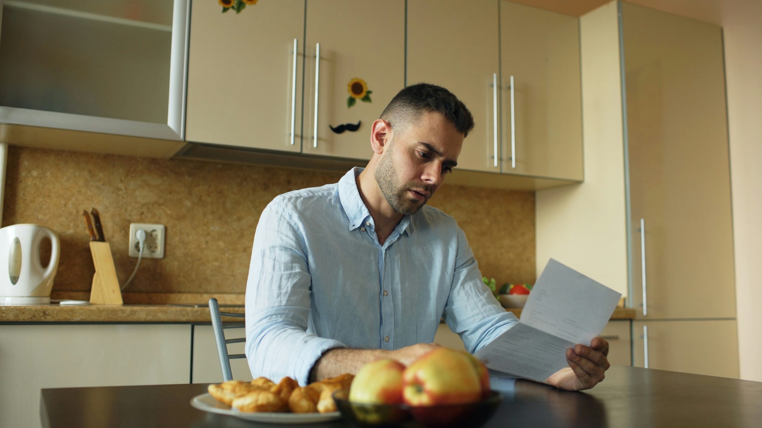 A man sits at a kitchen table reading a document with a serious, focused expression, holding papers in his hands while food sits untouched on the table, with light-colored cabinets, a kettle, and kitchen items in the background, suggesting concern or contemplation in a quiet home setting.